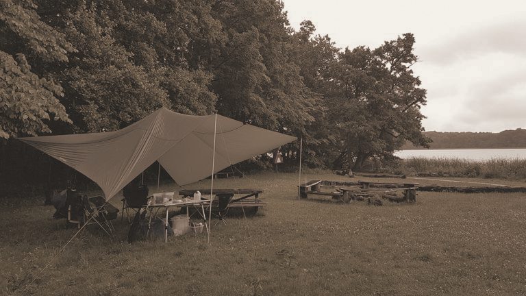 A green tarp pitched at the edge of a wood by a lake, a table of supplies and camp chairs beneath it, a log fire-circle nearby, in a warm-toned photograph.