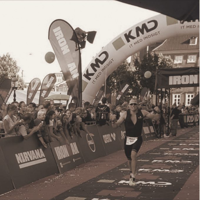 Running down the finish chute under the Ironman arch, arms wide, crowds along the barriers, in a warm-toned photograph.