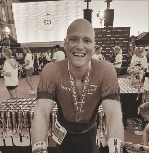 A smiling athlete leaning toward the camera, finisher medal around the neck, race village behind, in a warm-toned photograph.