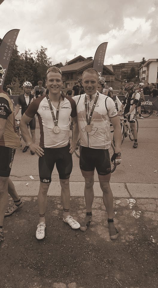 Two riders with finisher medals at the line, bikes and crowds behind, in a warm-toned photograph.