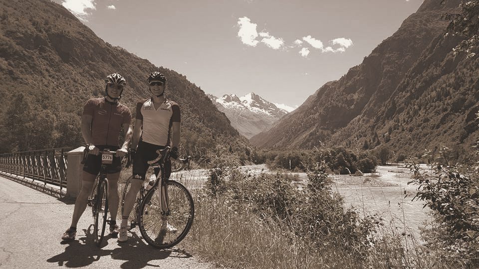 Two riders with race numbers pausing on a bridge over a glacial river, a snow-capped massif down the valley, in a warm-toned photograph.
