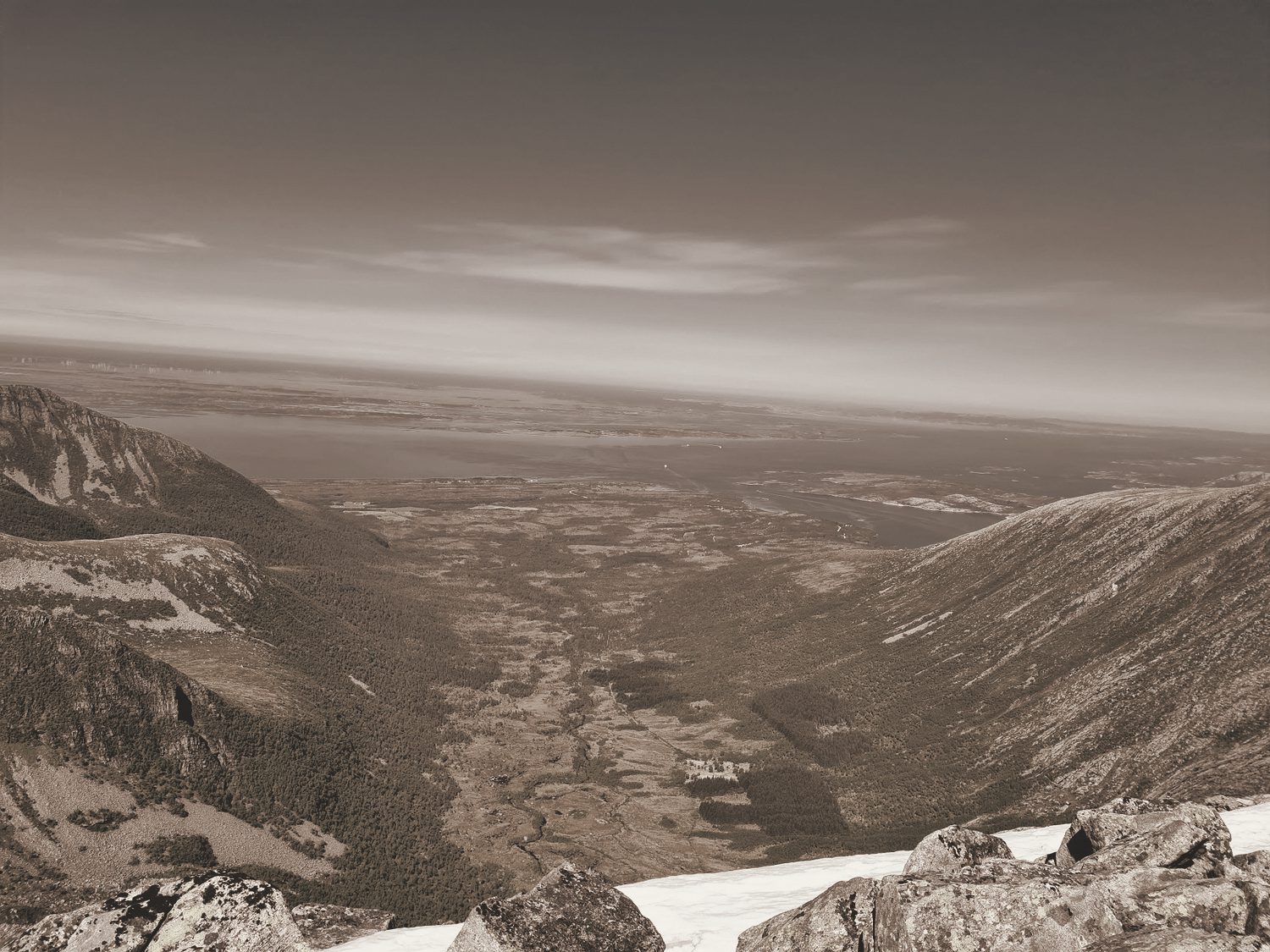 The view from a summit down a long green valley to a distant fjord, in a warm-toned photograph.
